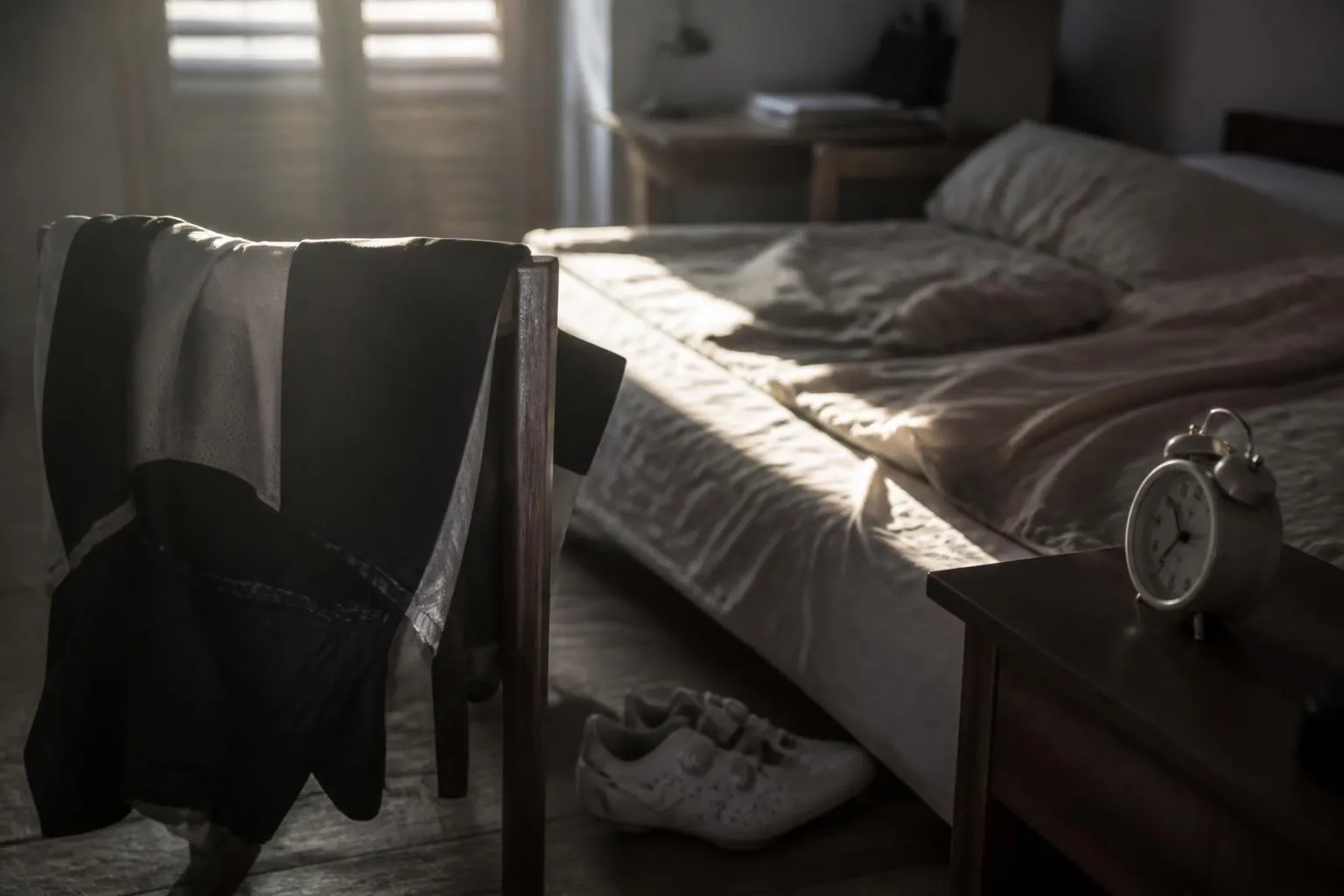 Une chambre aux volets mi-clos au petit jour, un lit défait, une tenue de cycliste pliée sur une chaise et des chaussures de route posées au sol